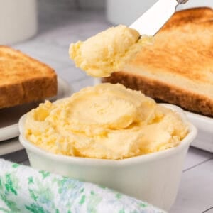 A knife spreads whipped butter from a white bowl, with toasted bread slices and a green-patterned cloth nearby on a light surface.