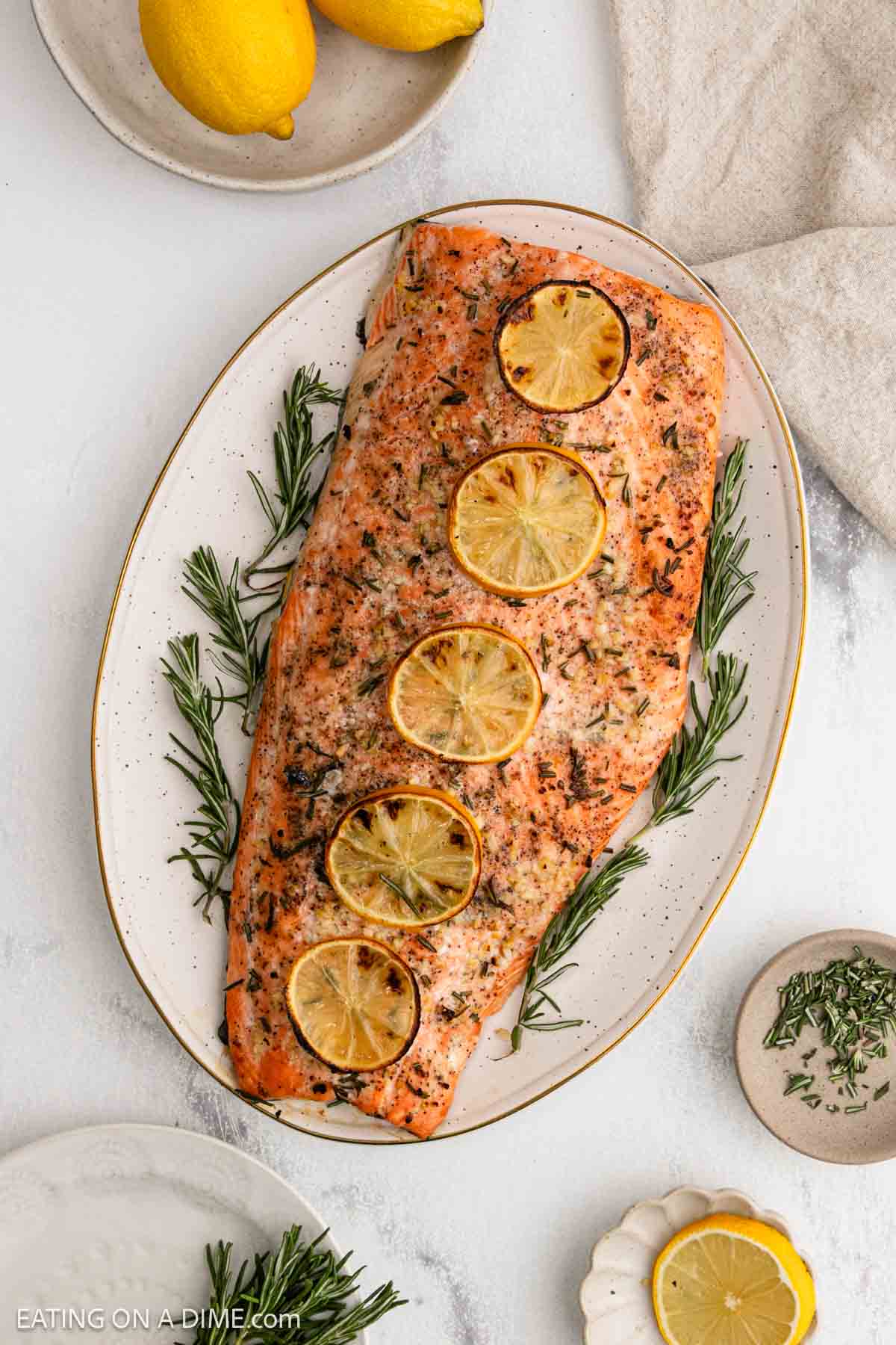 A baked salmon fillet garnished with four lemon slices and fresh rosemary sprigs is served on a white oval platter. Lemons, chopped herbs, and a beige cloth are nearby on the white surface.