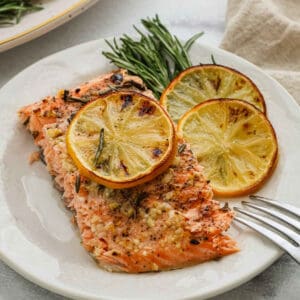 A plate with a baked salmon fillet topped with minced garlic and herbs, garnished with two slices of roasted lemon and fresh rosemary, with a fork resting nearby.
