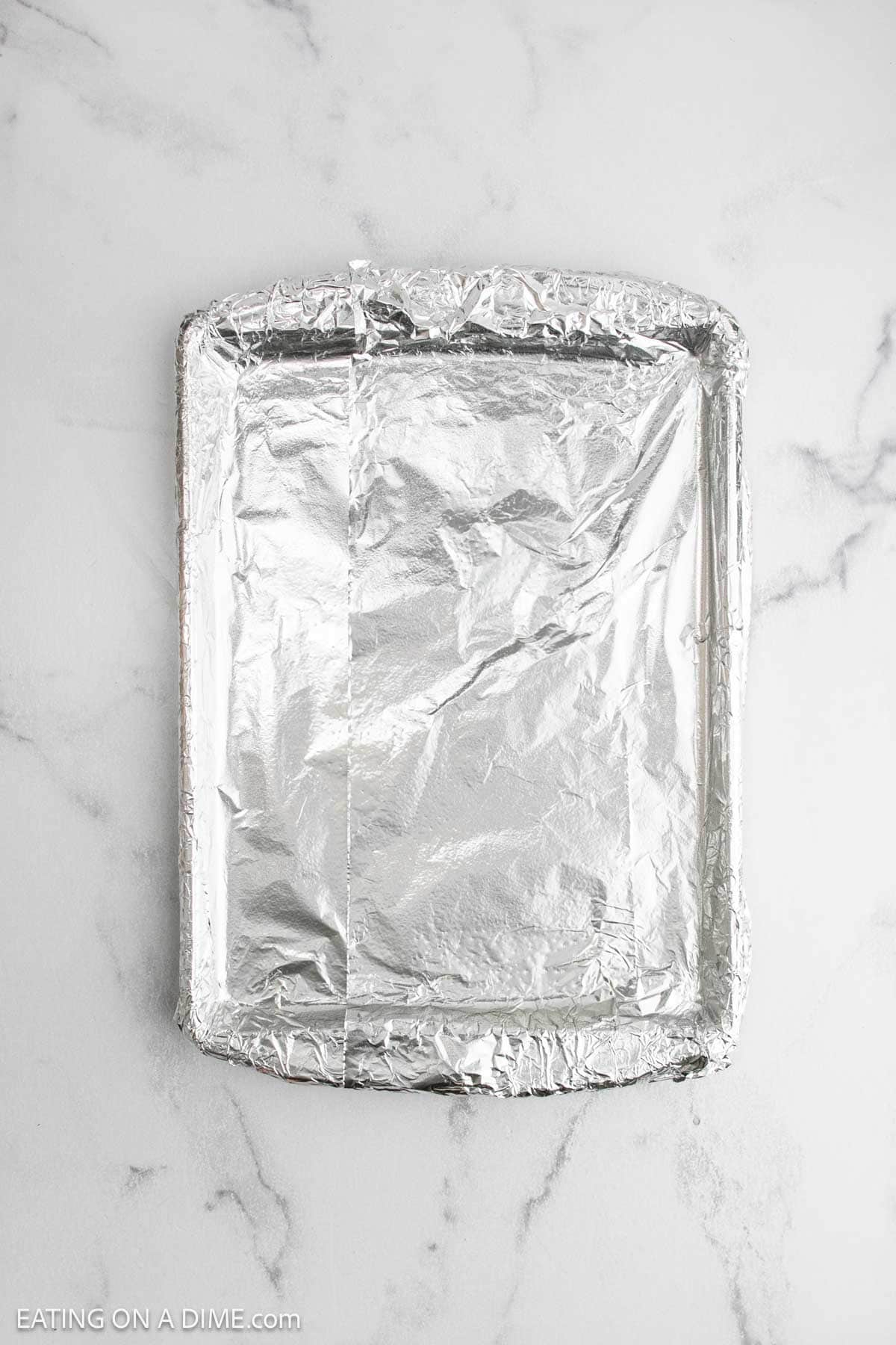 A rectangular baking pan lined neatly with aluminum foil, ready for baked salmon, sits on a white marble countertop.