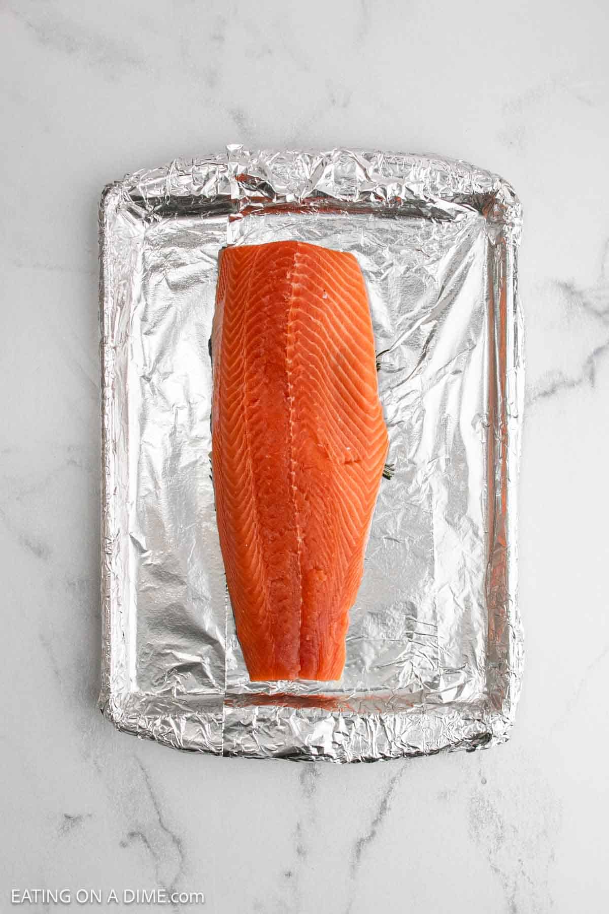A raw salmon fillet sits on a foil-lined baking tray, ready to become delicious baked salmon, placed on a light marble countertop.