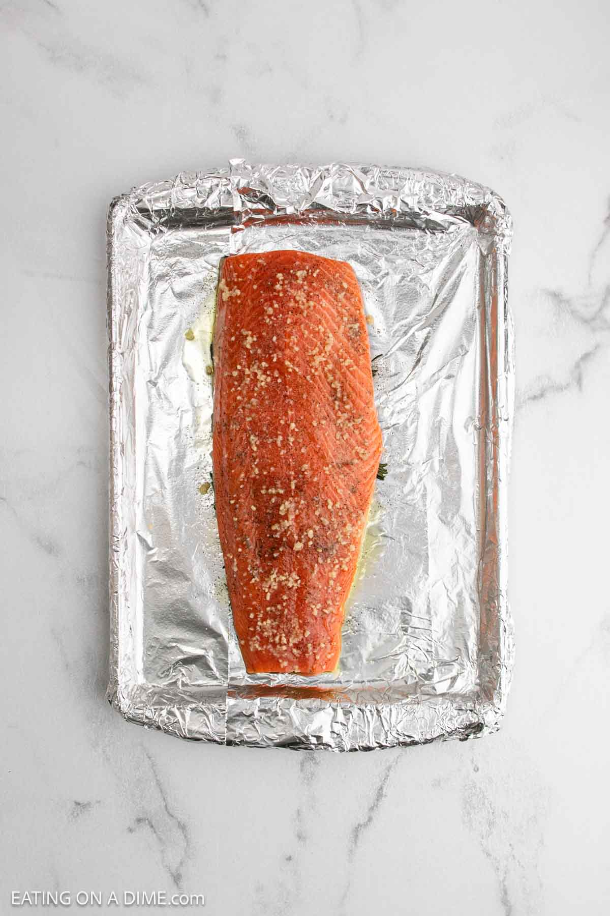A raw salmon fillet, perfect for baked salmon, is seasoned with spices and rests on a foil-lined baking sheet. The pan is placed on a white marble surface, ready for the oven.