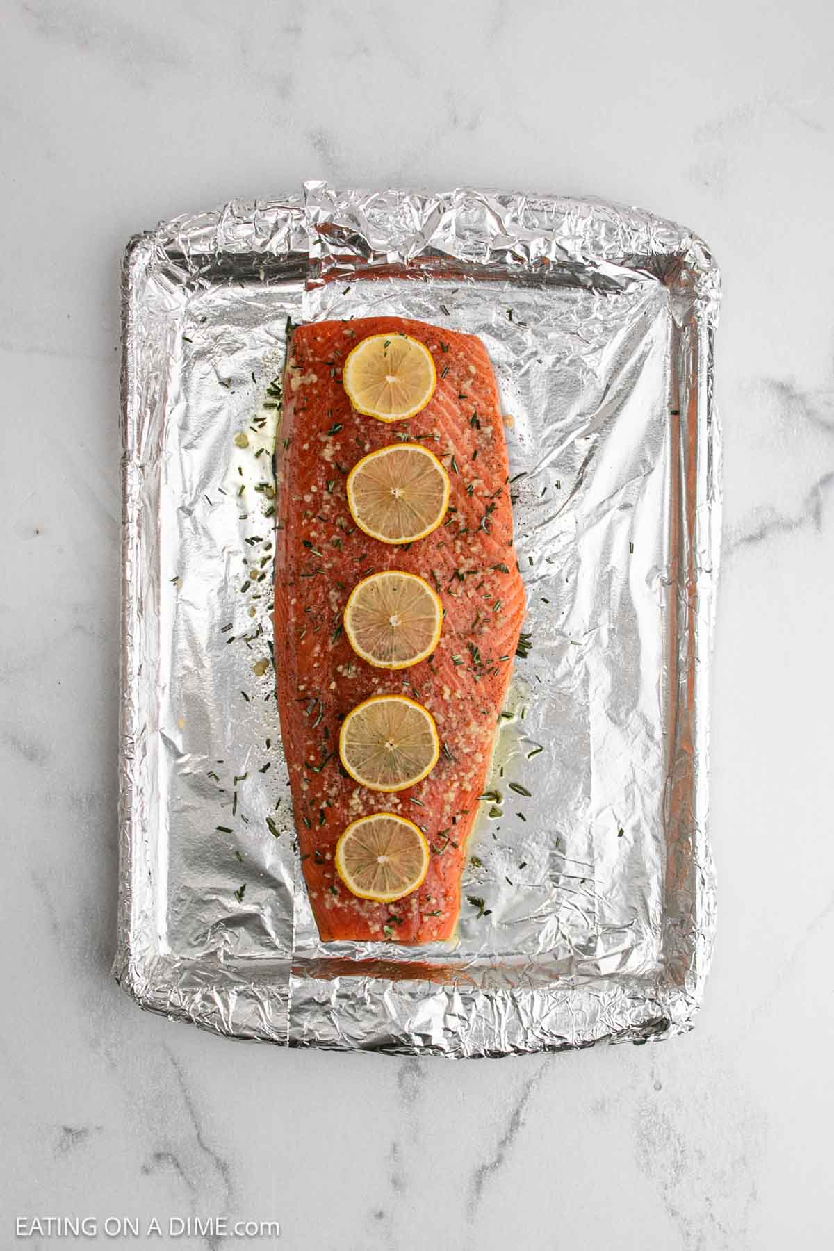 A raw salmon fillet topped with five lemon slices and herbs sits on a foil-lined baking sheet, ready to become delicious baked salmon.