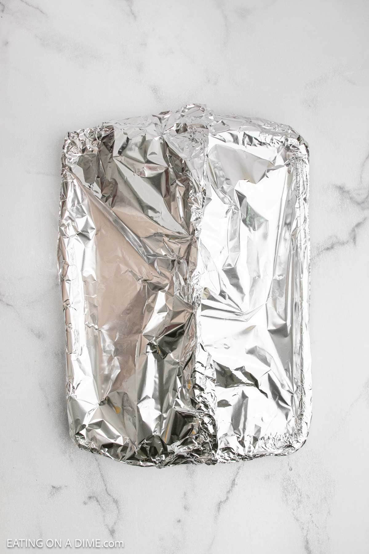 A rectangular baking dish, likely holding baked salmon, is covered completely with crinkled aluminum foil and sits on a white marble surface.