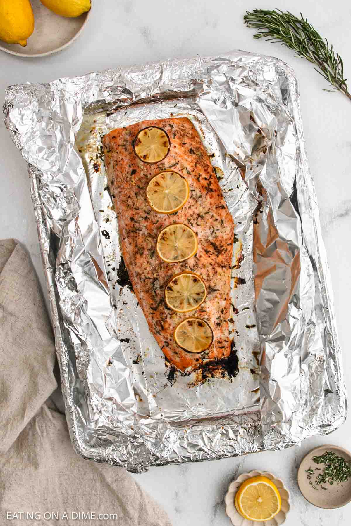 Baked Salmon fillet topped with five lemon slices, seasoned with herbs, and placed on a foil-lined baking sheet. Fresh rosemary, lemon, and a small bowl of seasoning are nearby on the marble surface.
