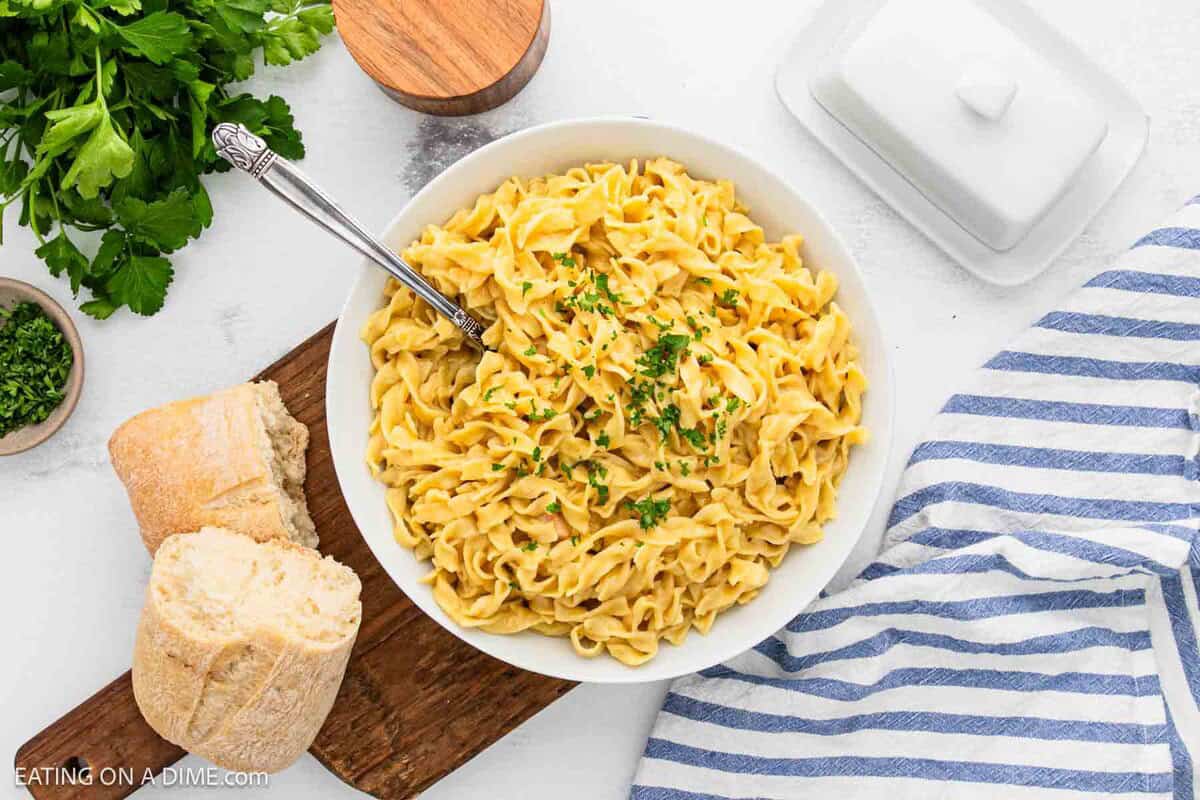 A bowl of creamy fettuccine Alfredo noodles garnished with chopped parsley, served with a piece of bread on a wooden board. A butter dish, fresh parsley, and a striped towel complete this inviting cafeteria-style setup on a white surface.