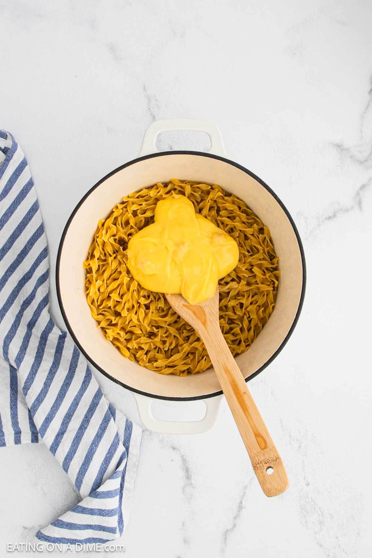A white pot filled with Cafeteria Noodles sits on a white surface. A wooden spoon holds a portion of melted cheese sauce above the noodles. A blue and white striped towel rests beside this nostalgic cafeteria food classic.