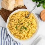 A bowl of creamy fettuccine alfredo noodles garnished with chopped parsley, served with a fork. Fresh bread and a striped napkin sit nearby on a light tabletop, capturing the comfort of a classic cafeteria meal.