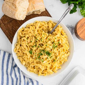 A bowl of creamy fettuccine alfredo noodles garnished with chopped parsley, served with a fork. Fresh bread and a striped napkin sit nearby on a light tabletop, capturing the comfort of a classic cafeteria meal.