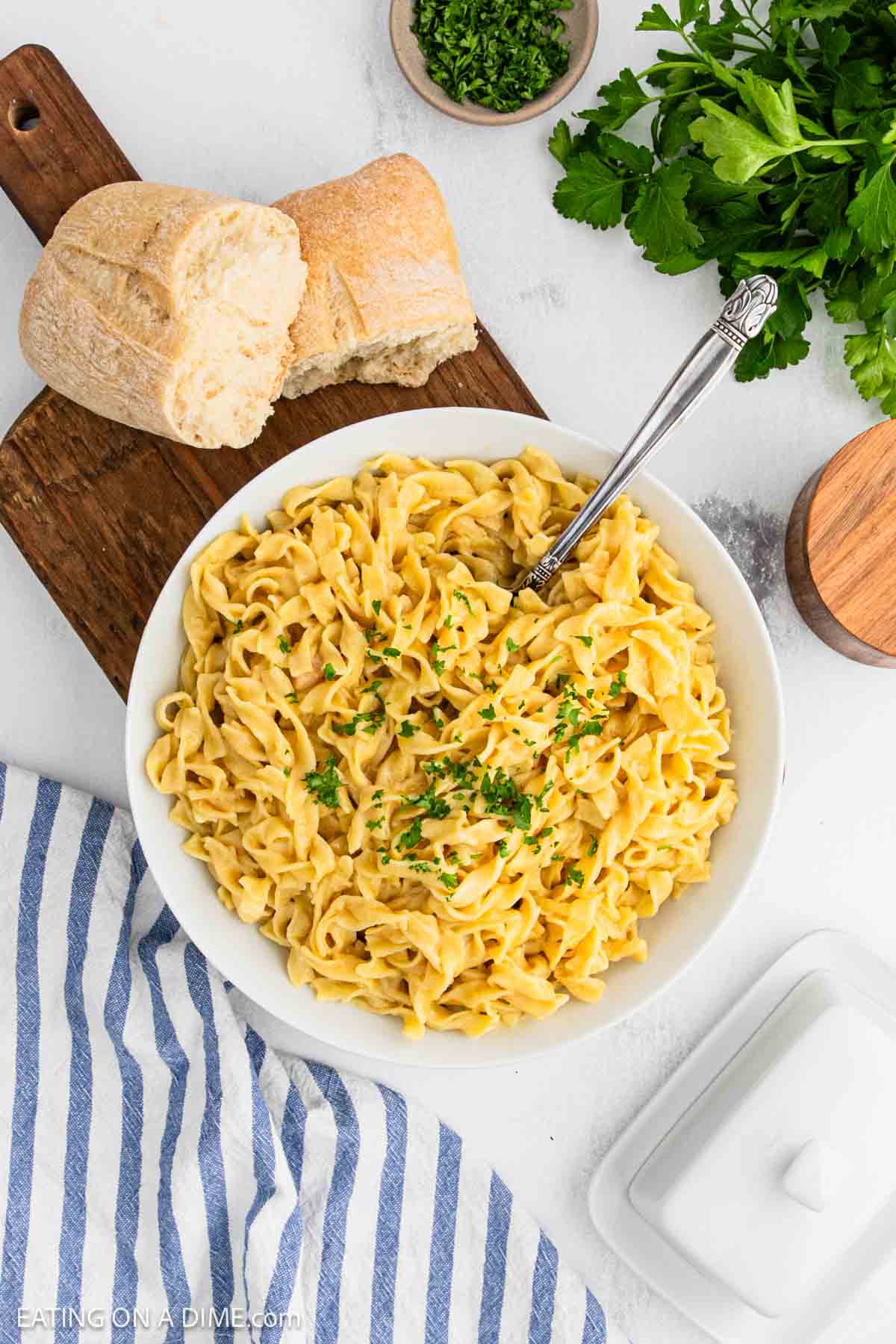 A bowl of creamy fettuccine, reminiscent of classic Cafeteria Noodles, garnished with chopped parsley and served with a metal fork. Slices of crusty bread and fresh parsley rest on a wooden board beside a striped cloth napkin.