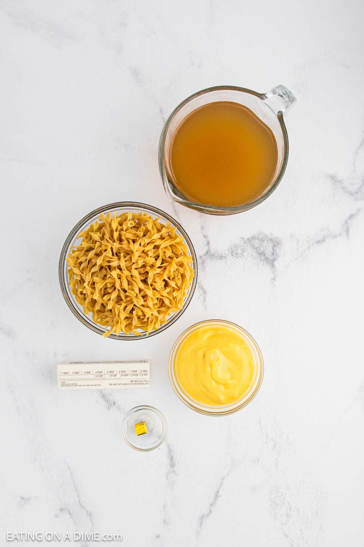 Overhead view of measured ingredients on a marble surface: cafeteria-style glass measuring cup with broth, a bowl of uncooked noodles, a bowl of creamy yellow soup, a stick of butter, and a bouillon cube.