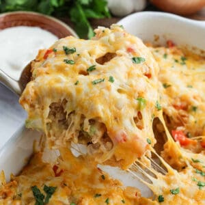 A cheesy, golden-baked Chicken Fajita Casserole is being scooped from a dish, with melted cheese stretching. Diced vegetables and herbs are visible, and a bowl of sour cream sits in the background.