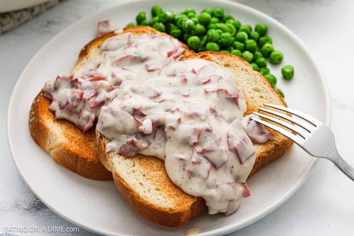 Two slices of toasted bread are topped with rich chipped beef gravy, served on a white plate alongside green peas and a fork.