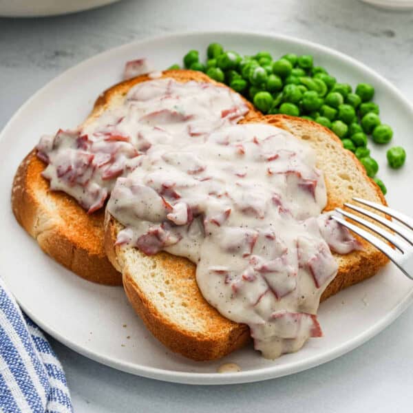 Two slices of toasted bread are topped with creamy chipped beef and gravy, served on a white plate with a side of green peas. A fork rests on the plate next to the chipped beef toast.