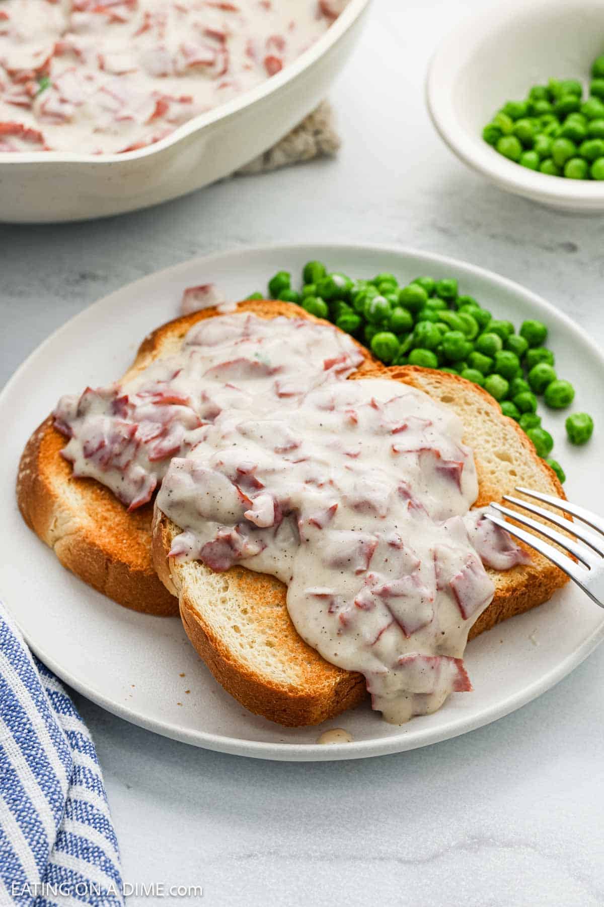 Two slices of toasted bread with creamy chipped beef gravy are served on a white plate alongside green peas. A fork rests nearby, and extra chipped beef gravy and peas can be seen in bowls in the background.