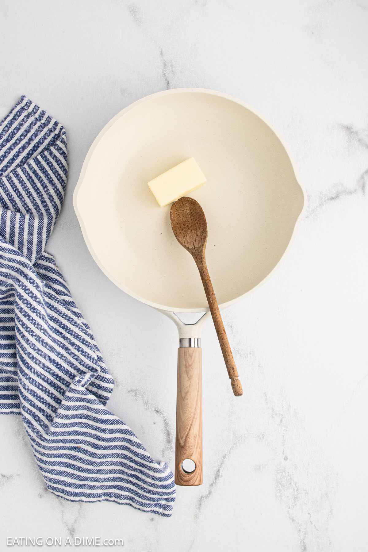 A white frying pan with a wooden handle holds a pat of butter and a wooden spoon, ready for making chipped beef. A blue and white striped kitchen towel is placed beside the pan on a white marble surface.