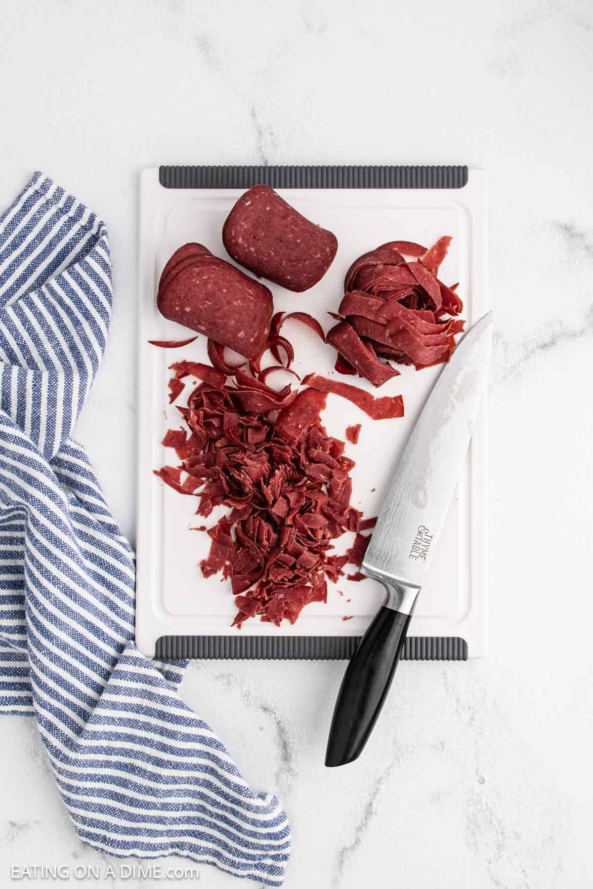 A white cutting board with sliced and chopped beef, perfect for beef recipes, a chef’s knife, and a blue-and-white striped kitchen towel on a white marble surface.