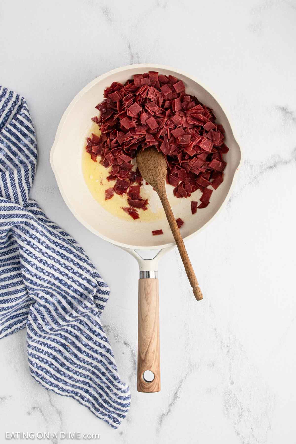 A white skillet with chopped corned beef and chipped beef, melted butter, and a wooden spoon sits on a marble countertop next to a blue and white striped kitchen towel.