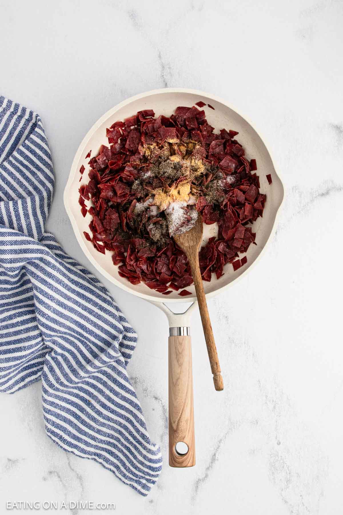 A white skillet filled with chopped chipped beef, seasonings, and a wooden spoon sits on a marble countertop next to a blue and white striped kitchen towel.