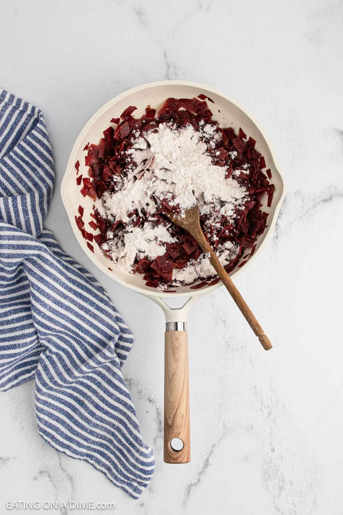 A skillet with chopped cooked beets and flour, being stirred with a wooden spoon, sits on a white marble surface next to chipped beef and a blue and white striped kitchen towel.
