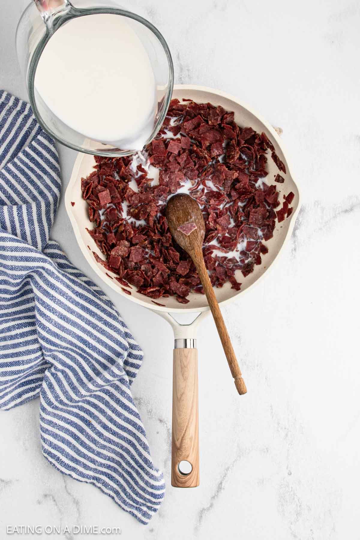 A skillet with chopped cooked dried beef and milk being poured in, a wooden spoon inside, and a blue-striped towel beside it on a marble countertop.