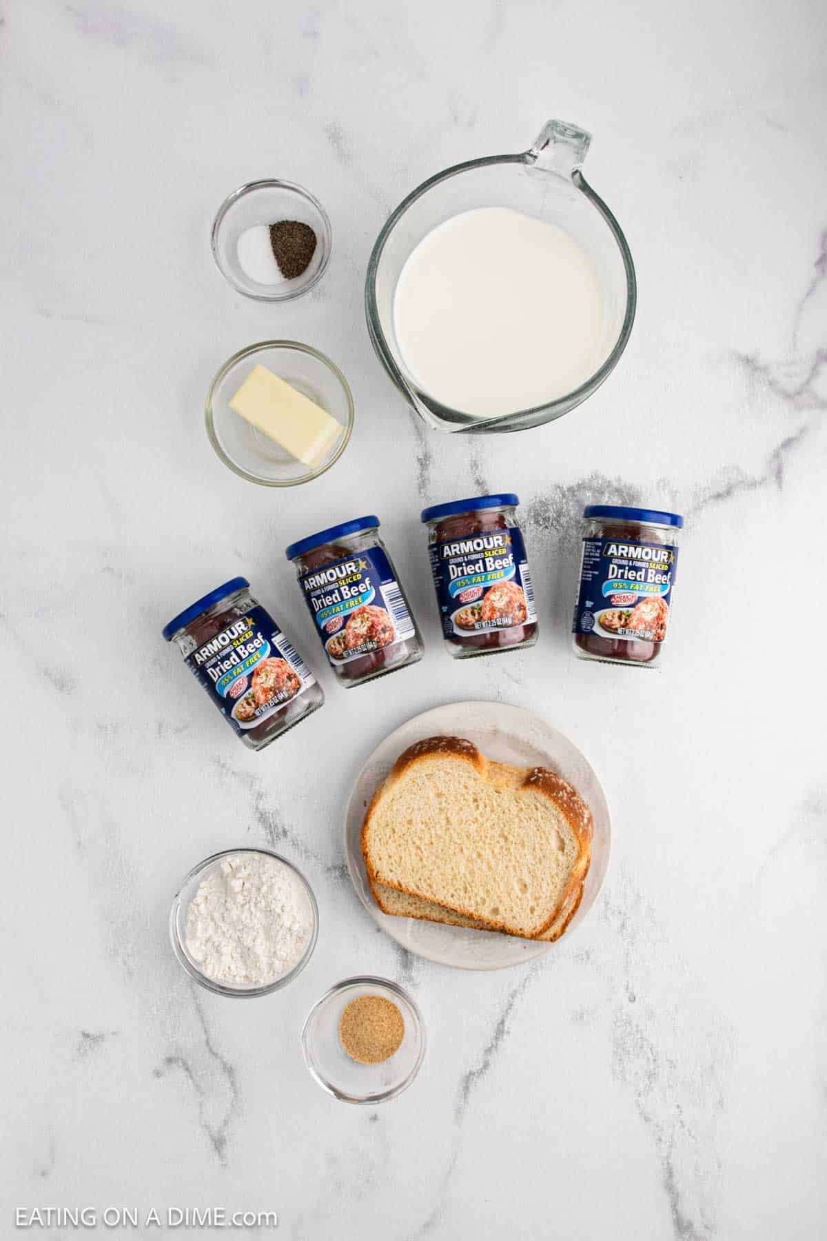 Ingredients for a Chipped Beef recipe are arranged on a white surface, featuring a glass measuring cup of milk, butter, salt, pepper, three jars of chipped beef, flour, bread slices, and a small bowl of brown seasoning.