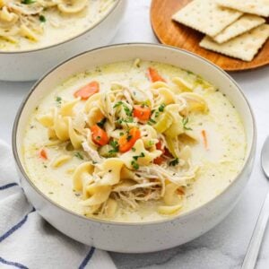 A bowl of creamy chicken noodle soup with egg noodles, shredded chicken, carrots, and celery, garnished with parsley. A plate of saltine crackers sits in the backgroundโperfect comfort for any Meal Plan 92.