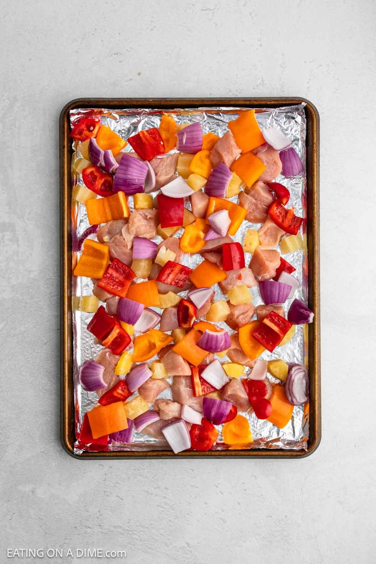 A baking sheet lined with foil is topped with chopped raw chicken, red onions, and assorted colorful bell peppers.