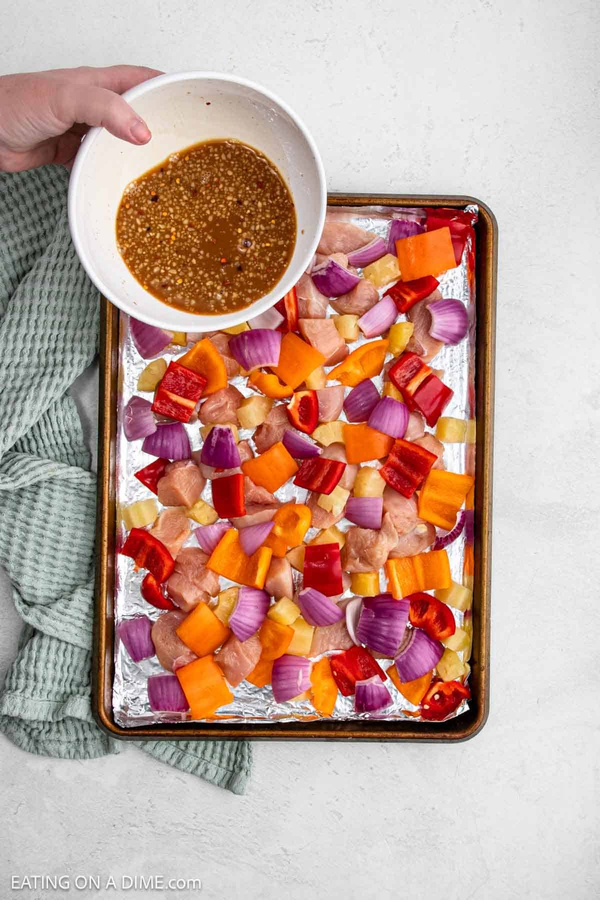 A hand pours marinade over a sheet pan filled with raw Chicken, red onions, bell peppers, and pineapple.