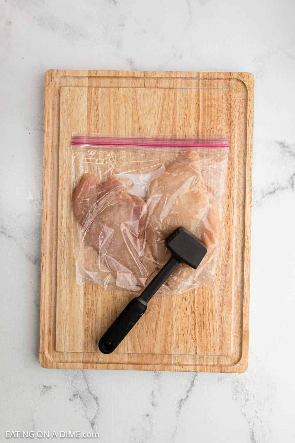 Raw chicken breasts in a plastic zip-top bag on a wooden cutting board with a meat tenderizer, ready to be prepped for Lemon Chicken Orzo, set on a light-colored countertop.