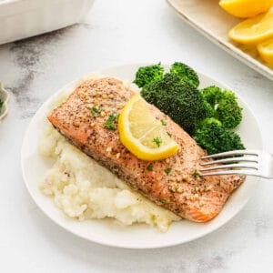 A plate of Lemon Garlic Salmon topped with a lemon slice, served over mashed potatoes and accompanied by steamed broccoli. A fork rests on the salmon.