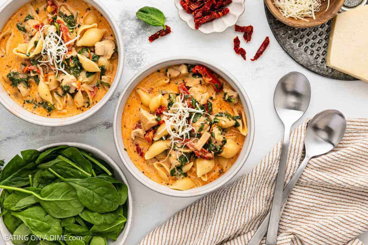 Two bowls of creamy Marry Me Chicken Soup with shell noodles, spinach, sun-dried tomatoes, and shredded cheese, surrounded by fresh spinach, spoons, a striped napkin, grated cheese, and sun-dried tomatoes on a white surface—an easy dinner delight.