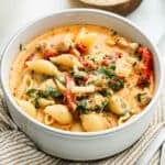 A white bowl filled with creamy pasta shells, chunks of Marry Me Chicken Soup-inspired chicken, sun-dried tomatoes, spinach, and herbs, set on a striped cloth with a slice of bread in the background.