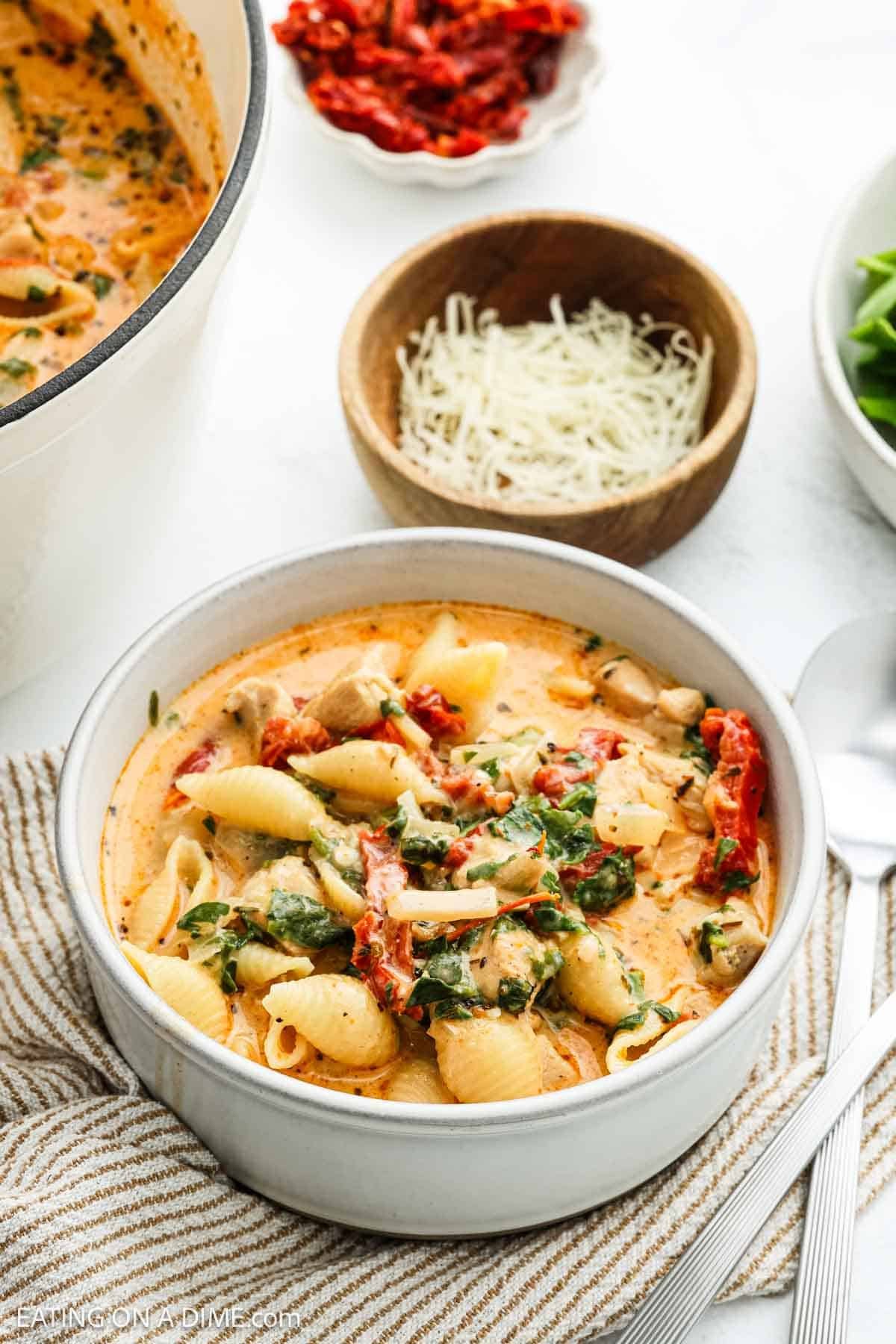 A bowl of creamy Marry Me Chicken Soup with shell noodles, spinach, sun-dried tomatoes, and chicken, garnished with herbs. Grated cheese and sun-dried tomatoes are in small bowls nearby on a white table.