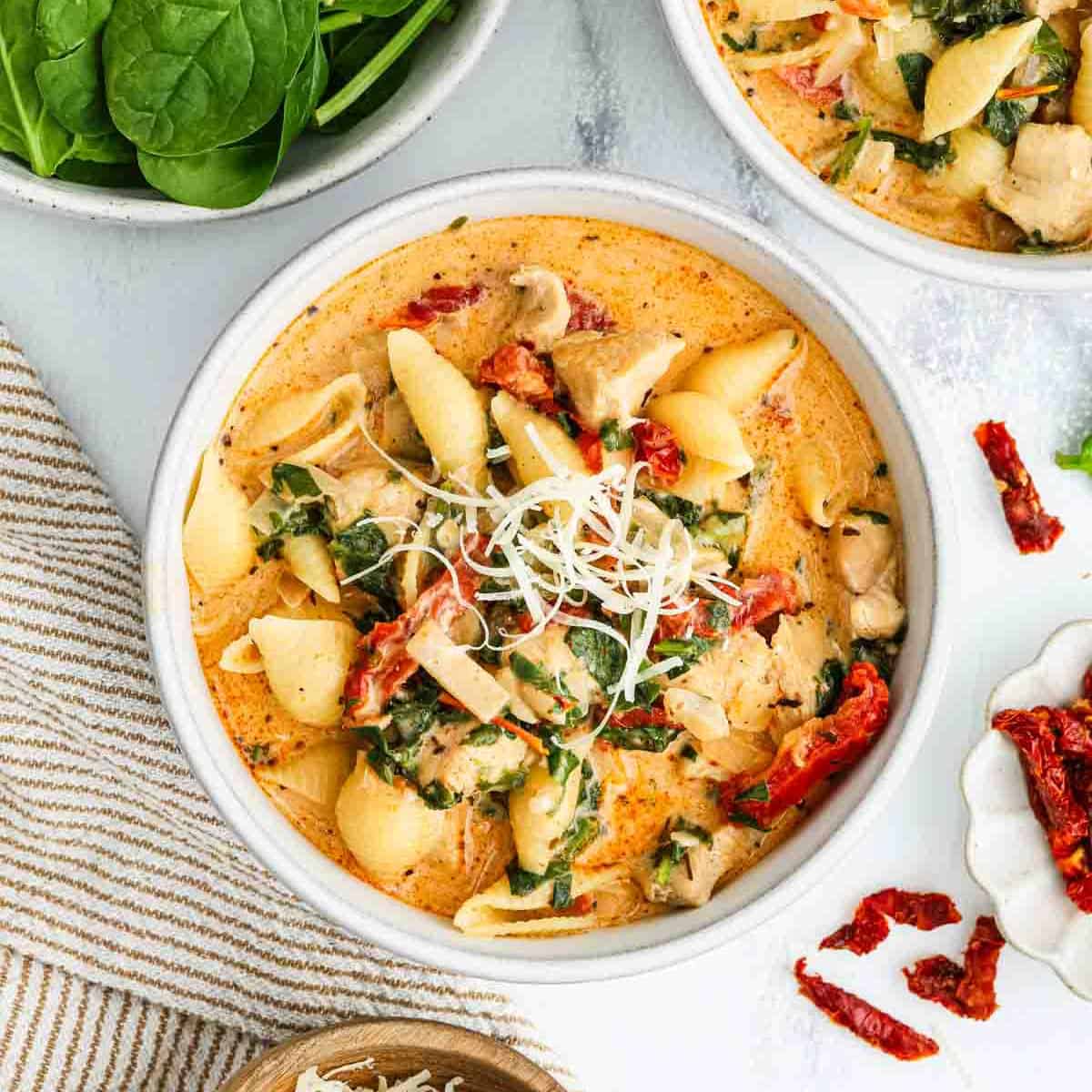 A bowl of creamy Marry Me Chicken Soup with pasta shells, shredded cheese, spinach, sun-dried tomatoes, and chunks of chicken, garnished with grated cheese, sits beside fresh spinach leaves and a striped napkin.
