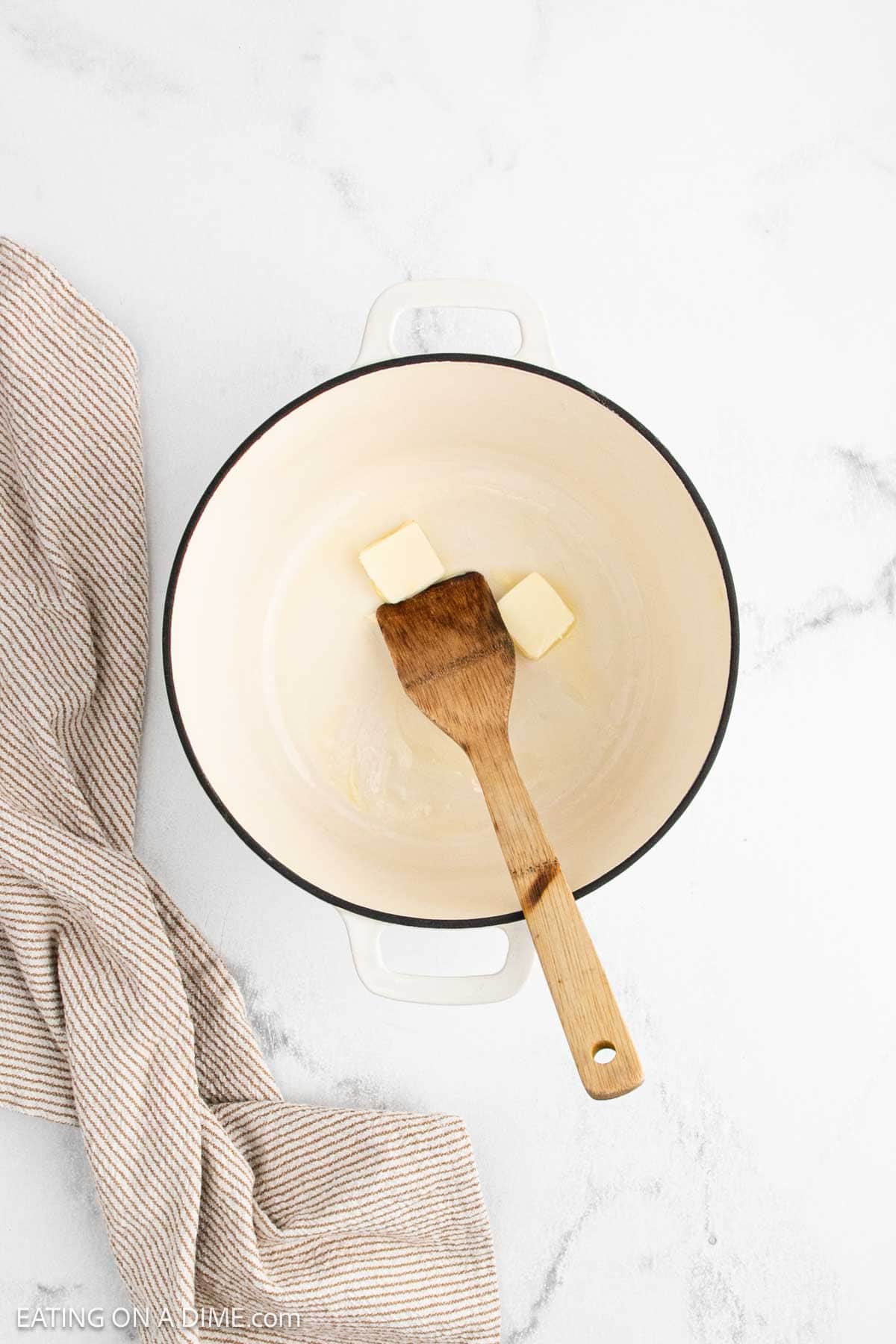 A white Dutch oven with two cubes of butter and a wooden spatula inside, ready to start Marry Me Chicken Soup, sits on a white marble surface next to a beige striped kitchen towel.