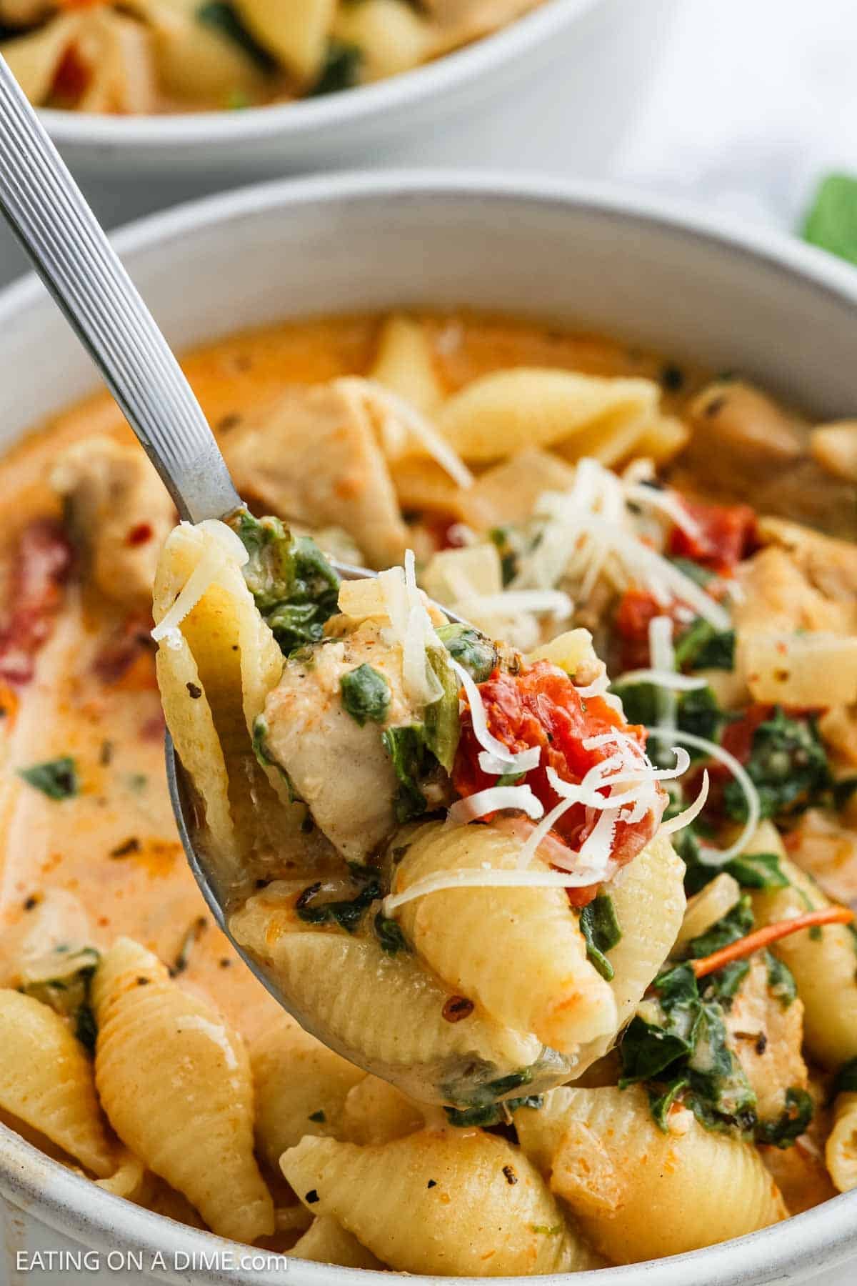 A spoonful of creamy pasta shells with chunks of chicken, spinach, tomatoes, and grated cheese held over a bowl of Marry Me Chicken Soup-inspired pasta dish.