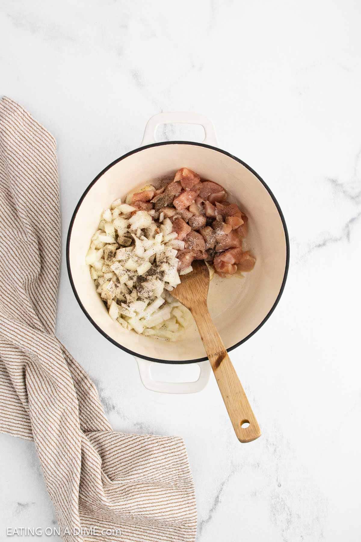 A white pot with chopped raw chicken, diced onions, and black pepper—perfect for Marry Me Chicken Soup—is being stirred with a wooden spoon on a marble surface next to a beige striped kitchen towel.