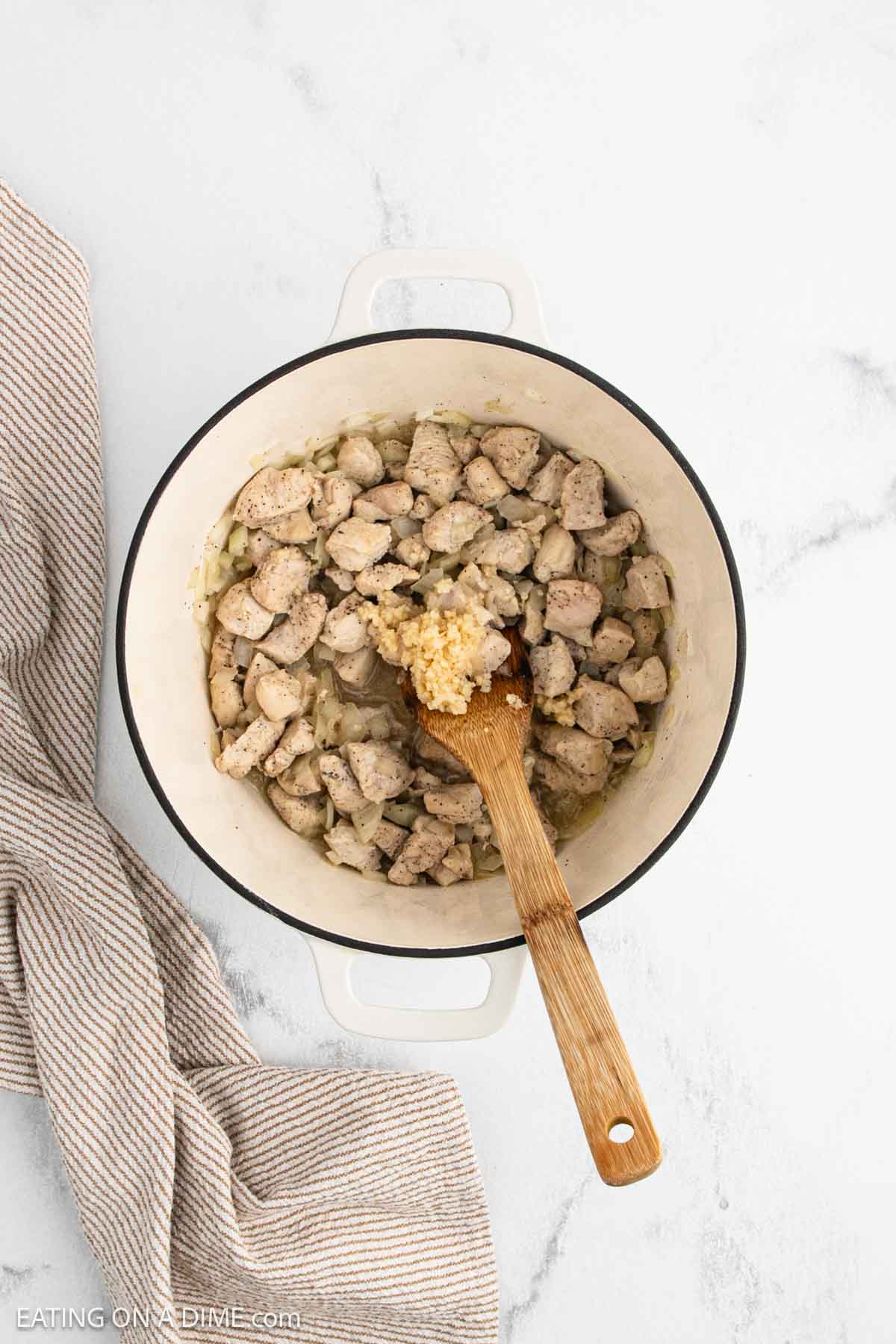 Cubed chicken pieces cooking in a white pot with minced garlic and onions, stirred with a wooden spoon—the start of a cozy Marry Me Chicken Soup. A beige striped kitchen towel sits next to the pot on a white marble surface.
