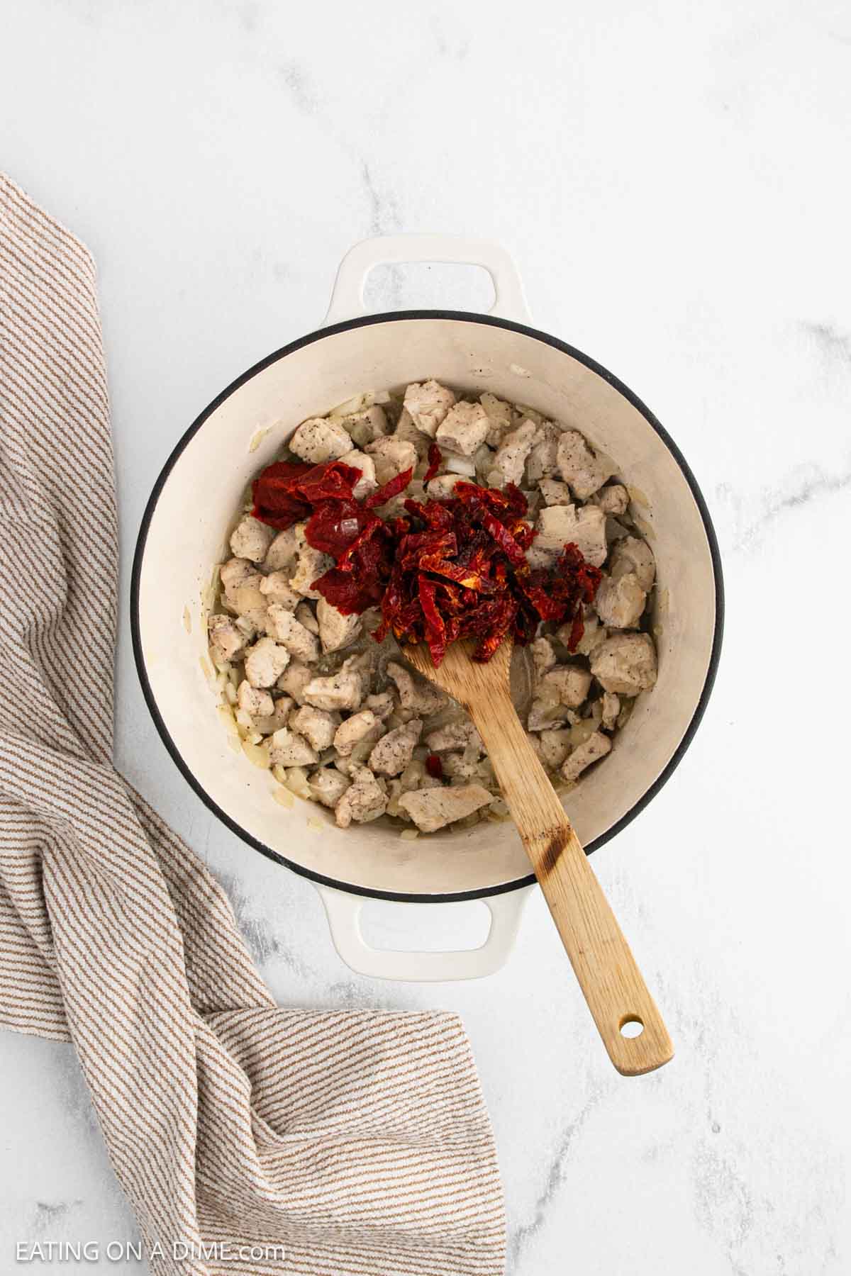 A white pot containing cooked diced chicken, onions, and a pile of tomato paste being stirred with a wooden spoon sits on a light marble surface—perfect for starting your own Marry Me Chicken Soup—next to a beige-striped kitchen towel.