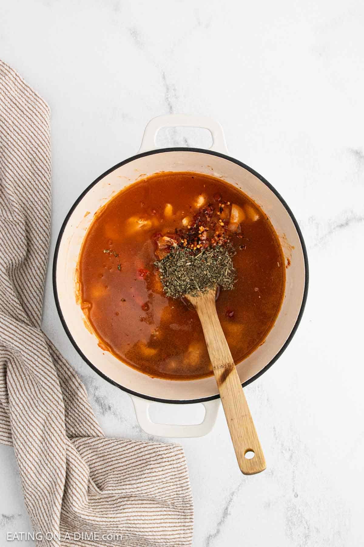 A white pot filled with Marry Me Chicken Soup, brimming with tomato-based broth, diced vegetables, and herbs, sits on a white marble surface. A wooden spoon rests in the pot, and a tan striped cloth is placed to the side.