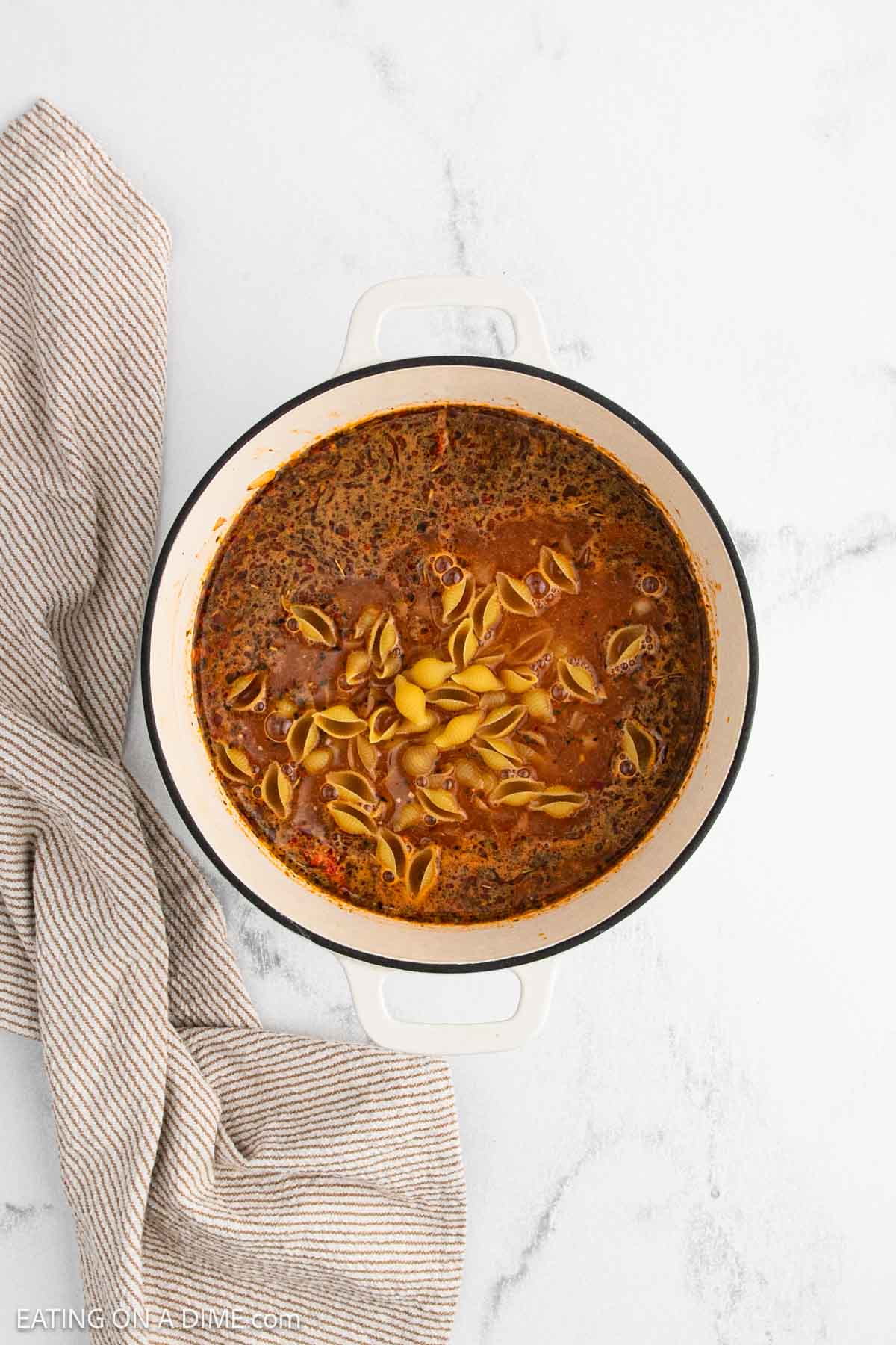 A white pot filled with Marry Me Chicken Soup featuring pasta shells and herbs sits on a marble surface next to a beige striped kitchen towel.