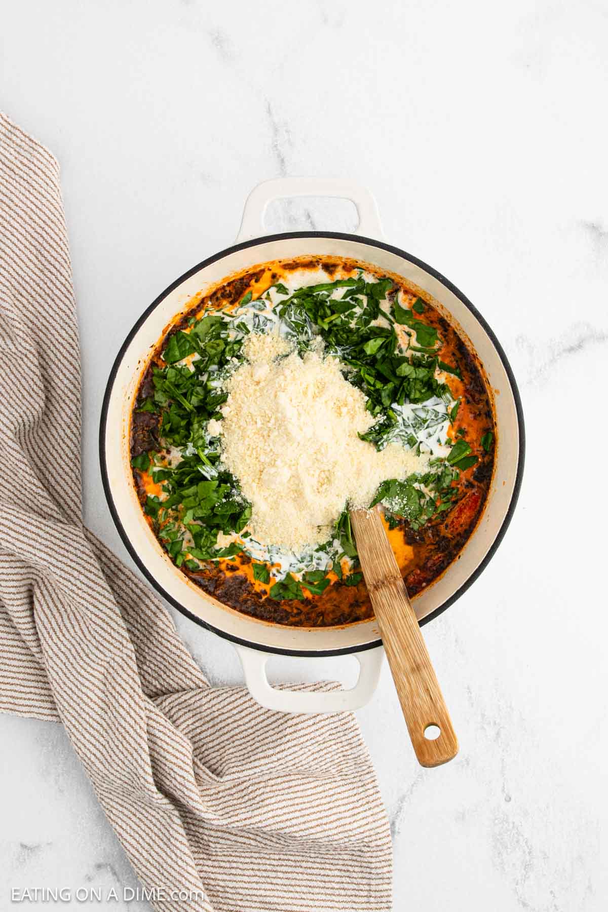A round white skillet with a creamy spinach and tomato mixture, reminiscent of Marry Me Chicken Soup, is topped with fresh chopped spinach and grated Parmesan cheese. It sits on a marble surface beside a beige striped kitchen towel; a wooden spoon rests in the pan.