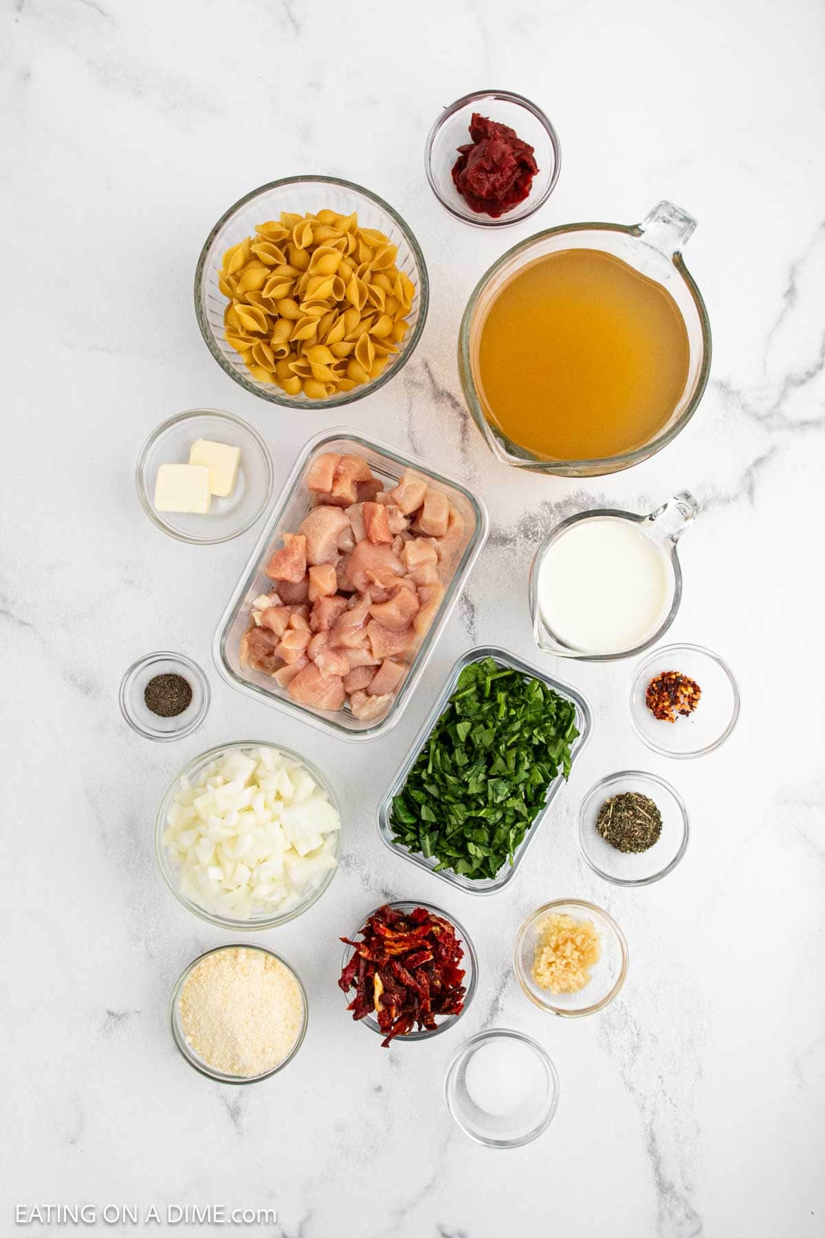 Various ingredients are arranged on a white surface, ready to create a creamy chicken soup, including dry pasta shells, chicken broth, diced chicken, chopped spinach, cream, diced onions, shredded cheese, butter, tomato paste, garlic and spices.