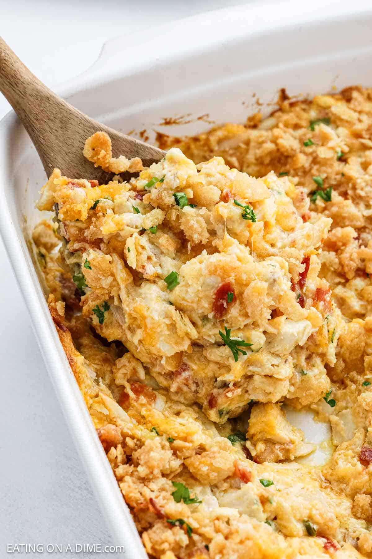 A wooden spoon scoops a cheesy, golden-brown Neiman Marcus Chicken Casserole with a crumbly topping from a white baking dish. Bits of herbs and diced vegetables are visible in the creamy filling.