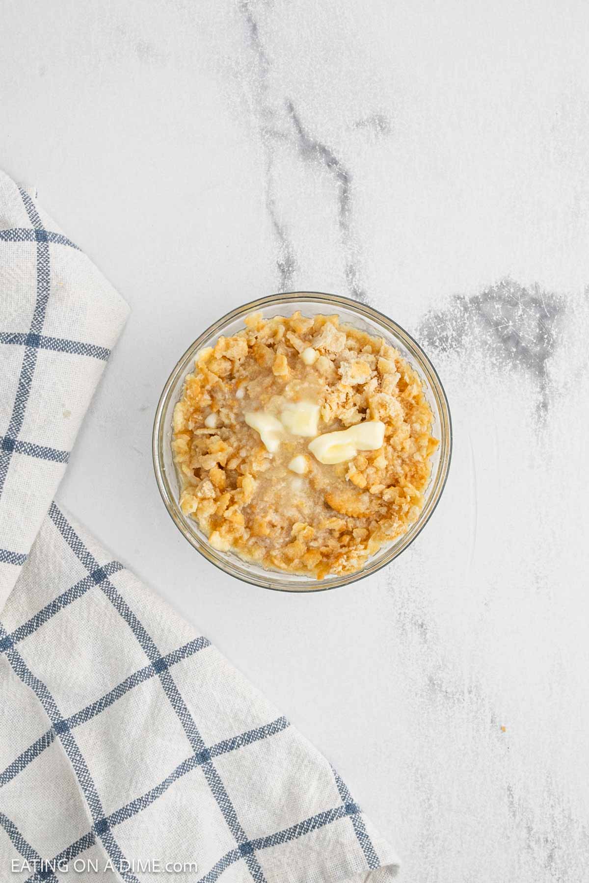A bowl of creamy oatmeal topped with melted butter and crumbled crackers, reminiscent of a comforting Neiman Marcus Chicken Casserole, sits on a white surface beside a white and blue checkered cloth.