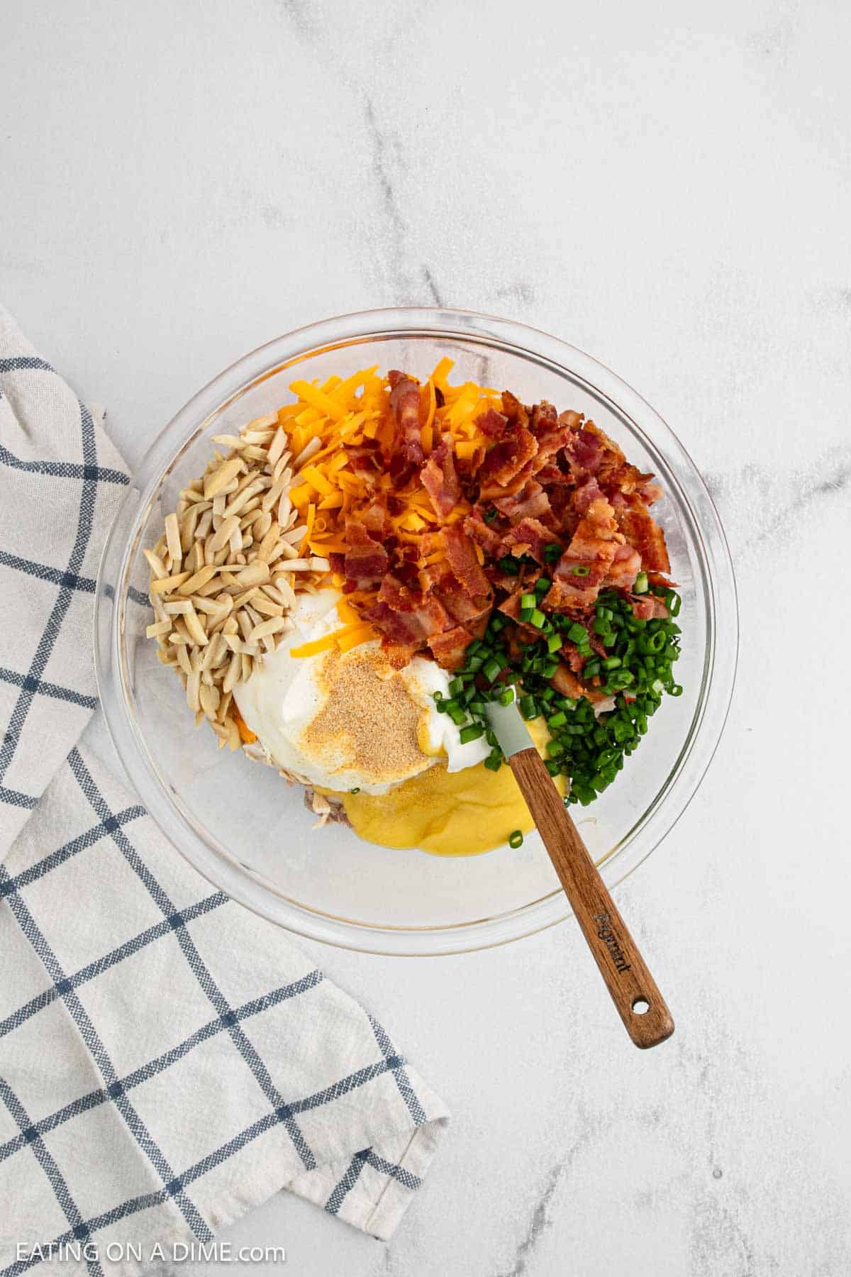 A glass bowl filled with Neiman Marcus Chicken Casserole ingredients like shredded cheddar cheese, cooked bacon, sliced almonds, green onions, mayonnaise, sour cream, garlic powder, and shredded chicken sits on a light surface near a checkered towel.