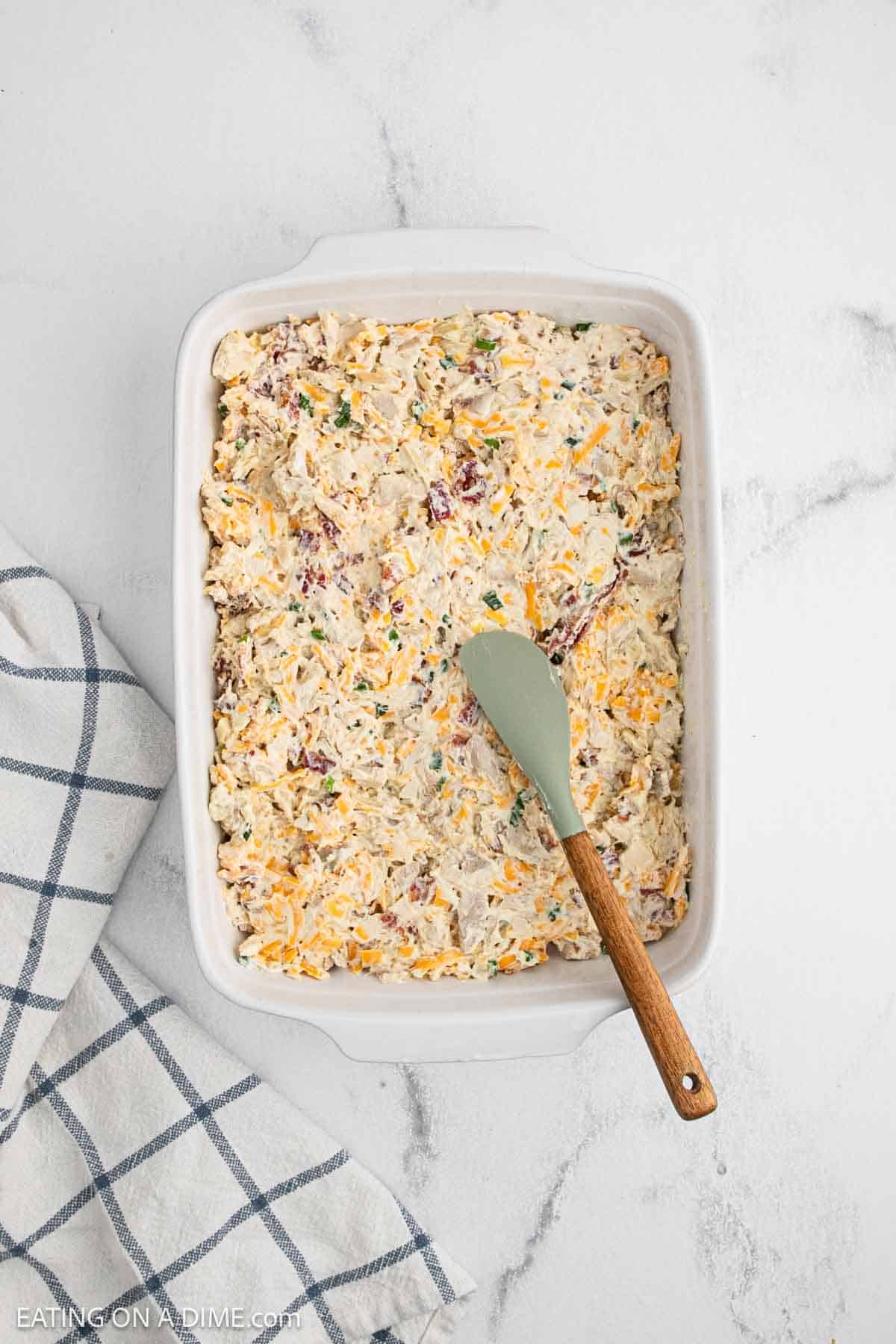 A white baking dish filled with an unbaked chicken casserole of shredded cheese, chicken, and herbs, with a green spatula resting inside. A white and blue checkered towel sits next to the dish on a marble surface.