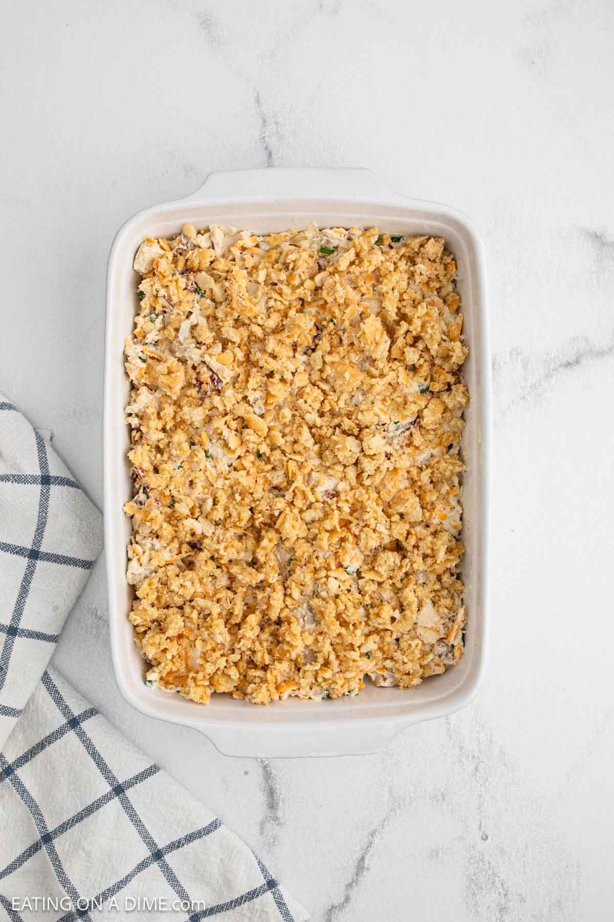 A white baking dish filled with a crumbly, golden Neiman Marcus Chicken Casserole sits on a white marble surface next to a blue and white checkered kitchen towel.