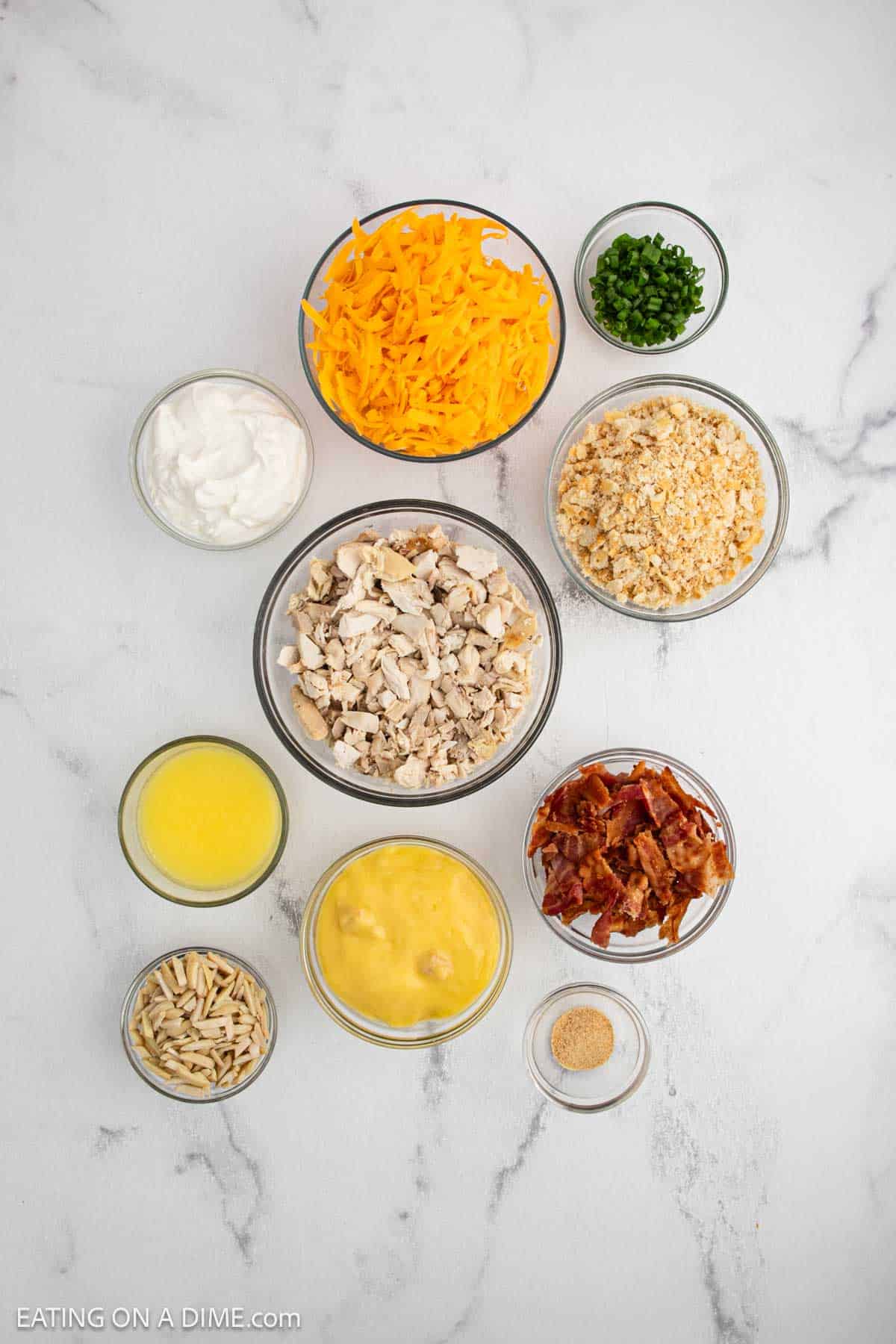 Nine bowls with Neiman Marcus Chicken Casserole ingredients on a white surface: shredded cheddar, green onions, sour cream, crushed crackers, chopped chicken, bacon pieces, sliced almonds, melted butter, cream soup, and garlic powder.